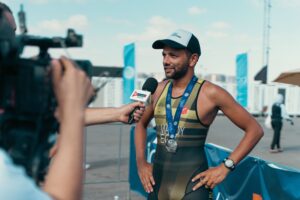 A medal-wearing triathlete speaks to a reporter holding a microphone after a race.