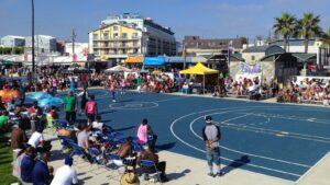 Players and spectators gather around a game on the blue basketball courts at Venice Beach in Los Angeles